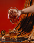 Hand pouring amber liquid from a glass into another glass on a draped fabric background