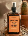 Room spray bottle labeled 'Small Town Rodeo' on a wooden block with a cactus in the background.