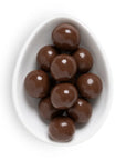 A top-down view of glossy brown chocolate-covered round candies piled in a white egg-shaped dish on a white background.