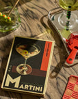 Cocktail setup with a Martini book, glasses, and bottles on a wooden surface.