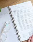 Open notebook with handwritten notes on a wooden surface, accompanied by glasses and a hand.