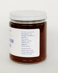 A glass jar filled with dark amber honey, featuring a white lid and a white label with blue text, set against a plain white background.