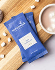 Two blue Moonstruck cocoa packets on a wooden board with a cup of hot chocolate and marshmallows.