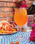 An orange-colored sparkling beverage in a wine glass, set on a blue mosaic tablecloth next to a plate of sliced citrus fruits, a bottle in an ice bucket, and bright pink flowers against a brick wall background.