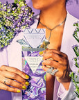 A person in a white blazer holds a lavender bar package from Compacts de Parfums, surrounded by purple and green flowers against a lavender background.