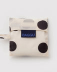 A folded reusable bag from BAGGU with a cream base, large black polka dots, and a navy blue brand label on the front.