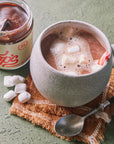 Ceramic mug with hot chocolate and marshmallows next to a jar of peppermint hot chocolate mix on a textured surface.