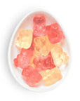 A top-down view of assorted gummy bear candies in pink, red, and orange-yellow colors, arranged inside a white, egg-shaped dish on a clean white background.