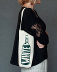 Person holding a tote bag with a wine bottle design and 'White Wine' text against a gray background