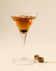 Martini glass with a brown cocktail and two cocktail cherries on a beige background