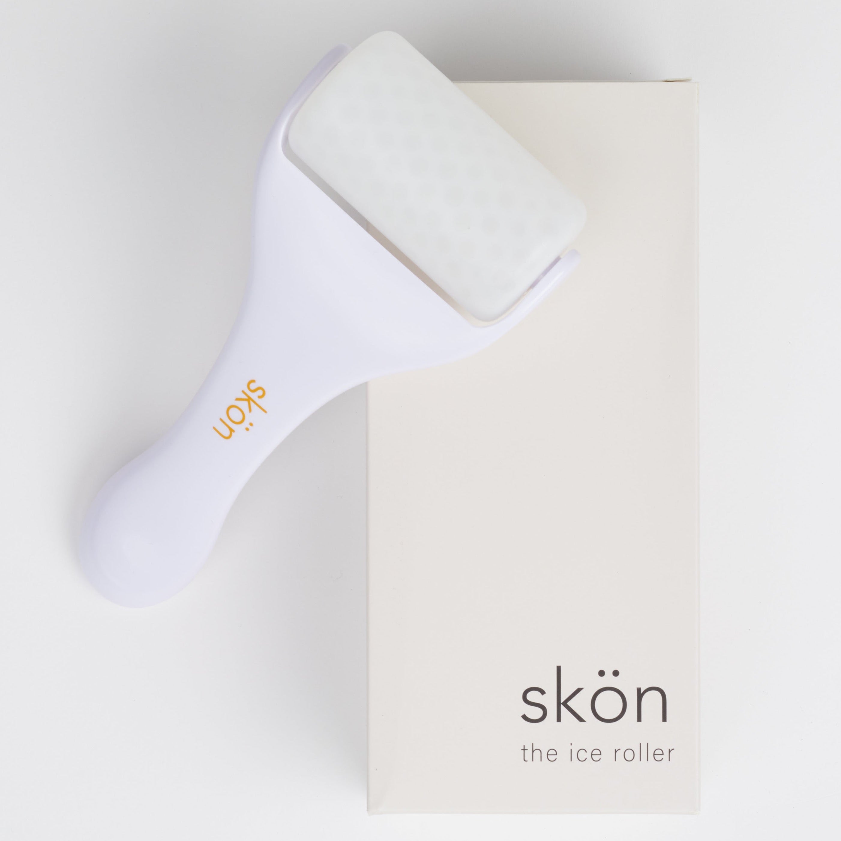A white ice roller with an tan-orange colored skön logo on the handle sitting atop a cream colored box that says &quot;skön the ice roller&quot; against a white background