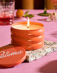 Candle with 'Paloma' branding on a pink surface with fruit and glasses in the background