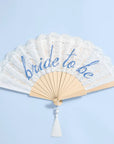 A folding hand fan with a cream lace overlay and blue script reading 'bride to be', attached to a light wood frame with a white tassel, set against a light blue background.