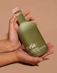 A person's hands holding a frosted olive green bottle of evie Organic Extra Virgin Olive Oil against a solid warm beige background.