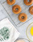 Six baked apple cider doughnuts rest on a metal cooling rack over a light marble surface. A white kitchen towel printed with green apple cider doughnut graphics and text lies nearby, with a glass bowl containing yellow liquid and another bowl partially visible at the bottom right.