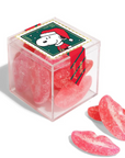 A clear square gift box filled with pink sugar-coated gummy candies, topped with a holiday-themed label featuring Snoopy in a Santa hat. Three lip-shaped gummies are placed in front on a white background.