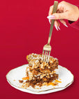 A hand with long pink nails holds a gold fork over a slice of layered dessert on a white plate with a gold rim, set against a solid red background.