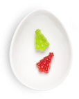 Two small Christmas tree-shaped gummy candies, one green and one red, displayed inside a white ceramic egg-shaped dish on a white background.