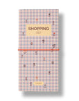 A long, narrow shopping list notebook with a pastel pink and blue gingham pattern, decorated with small floral illustrations. It features a beige label at the top reading 'SHOPPING list' and a bright orange elastic closure band across the middle, set against a plain white background with a soft drop shadow.