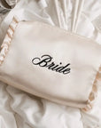 A cream satin bridal makeup bag with ruffled trim and the word 'Bride' embroidered in black script, displayed on a bed of white satin sheets.