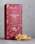 A red box of Grand Provisions Chocolate Chip bite size cookies with botanical illustrations, standing on a gray surface with two cookies in front.