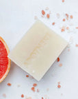 A rectangular bar of pale cream soap with the brand name 'ANOTHERE' embossed on it, styled on a white surface with scattered pink Himalayan salt crystals and a sliced grapefruit half visible on the left.