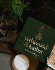 Box of 'cedarwood & leather' shower steamers by musee on a dark surface with a plant in the background.