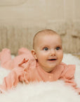 Baby in a pink outfit lying on a fluffy white rug with a wooden floor background