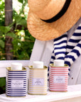 Three striped metal tins of olive oil are displayed on a white wooden table outdoors, with a person wearing a straw hat and a navy-and-white striped sweater partially visible in the background.