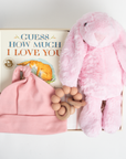 Pink baby hat, wooden teething ring, and plush bunny toy with 'Guess How Much I Love You' book on a light background