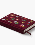 A hardcover notebook in deep burgundy velvet with colorful embroidered floral patterns on the cover, shown on a clean white background.