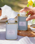 Person opening a striped can of Mevo+ basil olive oil on a table with a blurred outdoor background.