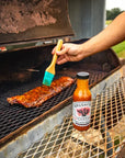 Person brushing ribs with a brush on a grill next to a bottle of Brushfire Farms sauce.