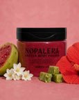 A jar of Nopalera Cactus Body Polish in Hibiscus scent, styled with a halved green guava, white jasmine flowers, green cactus pieces, and a red hibiscus flower on a pink background.