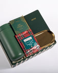 A curated gift set in a dark green striped box, featuring a dark green insulated bottle, a green notebook with gold lettering, a box of hot cocoa, a red tartan-patterned packet of Well-Apied hand sanitizer, and a gold-colored tray, all arranged on a white background.