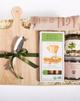 A gift hamper set on a white background, featuring a light wood cutting board with a green ribbon, a multi-tool, a box of Rustic Bakery organic sandwich bread, jars of California Native Mountain Wild Rose honey and olive oil, and a bag of Calamari.