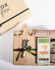 A curated gift box set from Box Fox, featuring a wooden cutting board with a green ribbon, a silver cheese knife, a package of Rustic Bakery crackers, and jars of gourmet spreads, arranged in shredded paper inside a kraft box next to the branded Box Fox gift box.