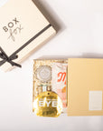 A curated gift box set from Box Box, featuring a cream-colored outer box with a black ribbon, and an open inner box containing a gold bottle of eye cream, a pink and white pouch, and a yellow-and-white striped box, all arranged on a white background.