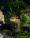 Green candle in a jar with a label, surrounded by ferns and rocks