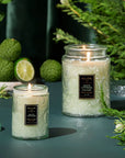 Two lit candles in glass jars with decorative labels on a dark surface with greenery.
