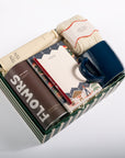 A curated gift box set containing a navy blue ceramic mug, a brown can of Flowers coffee, a cream tube of lotion, a patterned notebook, and a folded cream knit item, all arranged in a striped green and white box on a white background.