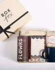 A curated gift box set from Box Fox on a clean white background, featuring a cream gift box with a black ribbon, a brown cylindrical tin labeled FLOWRS, a cream notebook with patterned borders, a navy blue ceramic mug, and a cream knit item wrapped in paper.