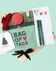 A curated gift set arranged on a light mint background, including a kraft gift box with shredded paper, a red hair claw clip, a round red soap bar labeled 'APOTHEKE', a mint green 'BAG OF TAGS' pouch, a white 'LIST MAKER' notepad, two green pens, and a white Christmas greeting card tied with a black ribbon.