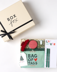 A Christmas-themed gift box set from 'Box fox' shown in a flat-lay view. The set includes a cream-colored gift box with a black ribbon, and an open box containing a mint green 'Bag of Tags' booklet, a red elastic hair tie, a pink circular item, a white 'List Maker' notepad, and two green pencils.
