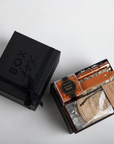 A black gift box with 'BOX box' printed on the lid sits beside an open box revealing a curated gift set with bath salts, a candle, and a packaged item on a light gray background.