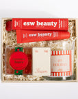 A curated gift set in a light wood box filled with shredded paper, featuring two red esw beauty lip balm tubes, a round red and green 'Champagne Bears' candle tin, a small cream-colored box with 'Tis The Season' script, and a striped red and white candle in a glass jar.