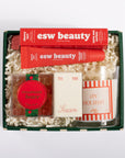 A curated gift box set containing an esw beauty lip balm tube, a red and green cherry lip balm, a candle, and a small card, all arranged in a dark green box filled with white shredded paper.