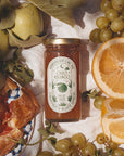 Jar of Oro Blanco grape jelly surrounded by fruits and pastries on a white cloth.