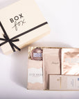 A luxury gift set from Box Fox, featuring a cream-colored box with a black ribbon next to an open box containing skincare and comfort items including Pearl Face Mask, Luxe Socks, and a chocolate bar, on a white background.
