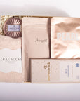 A curated gift box set containing a plush beige sweater, a round metallic rose gold soap, a package of Meriygold pouches, a package of Pearl Chocolate, and a box of Moonstruck Milk Chocolate, arranged in a box with shredded paper.